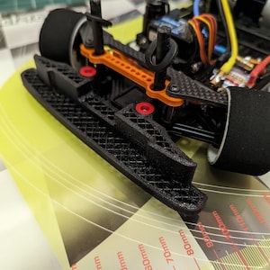 Puede incluir: Un chasis negro impreso en 3D para un coche de control remoto con brazos de suspensión naranja y negro. El chasis está sobre una superficie amarilla con una regla marcada en centímetros.