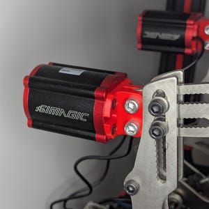 Könnte beinhalten: Nahaufnahme eines schwarzen und roten Elektromotors mit dem Text "#SIMMAGIC" auf der Seite. Der Motor ist an einer silbernen Metallhalterung mit zwei Schrauben befestigt.