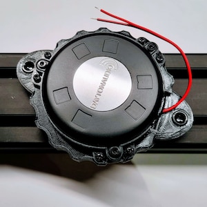 Puede incluir: Un transductor táctil Dayton Audio negro montado en una carcasa negra impresa en 3D con un borde festoneado. El dispositivo circular presenta un disco metálico plateado central con el logotipo de la marca y dos cables rojos expuestos.
