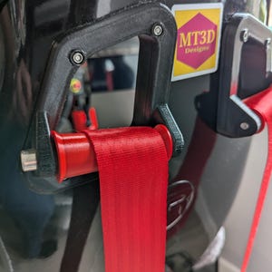 May include: A red racing harness strap is threaded through a black 3D printed part with a red roller. The part is attached to a black surface with a sticker that says "MT3D Designs".