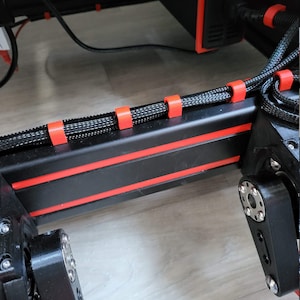 Può includere: Primo piano di un telaio di stampante 3D nero e rosso con cavi neri fissati da clip rosse. I componenti della stampante includono supporti neri e bulloni color argento. Lo sfondo è una superficie di legno di colore chiaro.