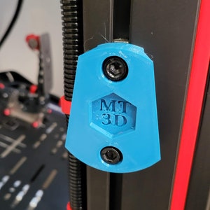 以下が含まれることがあります： 盾のような形をした明るい青色の3Dプリント製ブラケット。中央には六角形の切り抜きがあり、MT 3Dという文字が浮き彫りになっています。この部品は、上下2本の黒い六角ボルトで黒い垂直の金属レールに固定されています。