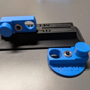 May include: Two blue 3D-printed components with circular openings and a small metal bearing. One component is attached to a black base with the text "MT3D". The other component is detached and has the text "MT3D" on it.