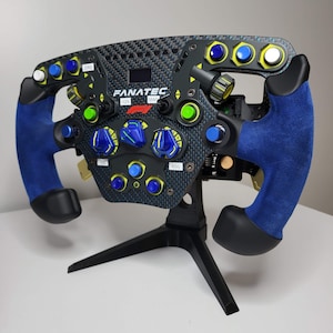Puede incluir: Un volante de carreras negro con detalles azules y amarillos. El volante tiene una variedad de botones e interruptores, incluida una pantalla. El volante está montado en un soporte negro.