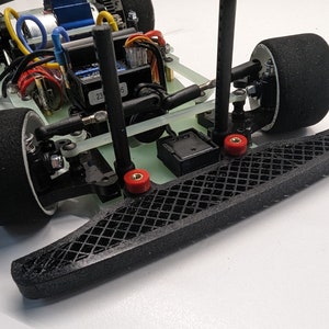 May include: A detailed view of a small RC car chassis. The model has black tyres, a black front bumper, and various electronic components. Red and black wires are visible, along with a blue component and a clear chassis.