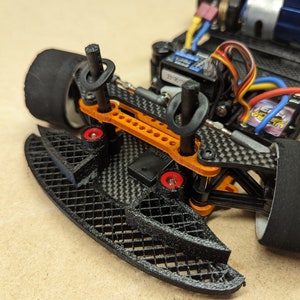 May include: A close-up of a black and orange RC car chassis with a carbon fibre body. The car has a black front bumper with a grid pattern and red and orange accents.