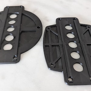 Puede incluir: Dos placas circulares de plástico negro con una serie de agujeros y una ranura vertical en el centro. Las placas están diseñadas para montar y asegurar objetos.
