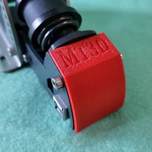 May include: A close-up of a red 3D printed part with the text "MT3D" embossed on the top. The part is attached to a black metal object.