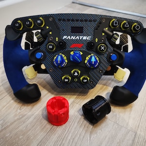 May include: A black racing steering wheel with blue accents and yellow buttons. The wheel has a carbon fiber design and the text "FANATEC" printed on it. The wheel is attached to a black and red adapter.