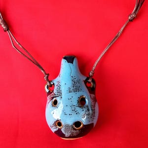 Può includere: Un ciondolo ocarina in ceramica blu con una collana di cordoncino marrone scuro. L'ocarina ha un design maculato e diversi fori. Lo sfondo è rosso pieno.