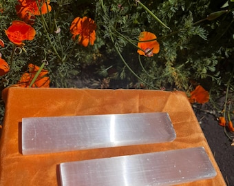 May include: Two rectangular, translucent selenite crystal wands are displayed on an orange surface, reflecting light. The wands are positioned horizontally, showcasing their smooth, polished surfaces. The background features vibrant orange flowers and green foliage.