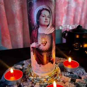 Può includere: Una candela cilindrica alta con un'immagine religiosa, circondata da tre lumini rossi accesi. La candela presenta un ritratto di donna con un'aureola e un cuore. La candela è posizionata su un centrino all'uncinetto con braccialetti di perline.