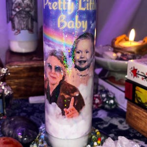 May include: A white prayer candle with the text "Pretty Little Baby" in gold lettering. The candle features a photo of a baby and an older woman. The background is a blue sky with a rainbow.