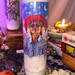 May include: A clear glass candle with a printed image of a person wearing a red hat and sunglasses, set against a blue sky with a crescent moon. The candle has a white wax base and a lit wick. Additional candles and crystals are in the background.