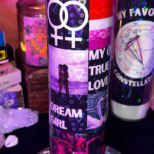 May include: A tall, pink candle with a decorative label featuring the text "MY OWN TRUE LOVE" and "DREAM GIRL". The label includes a lesbian pride flag design, hearts, and a couple embracing on a beach. A swan is also depicted.