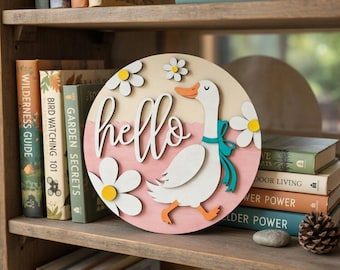 May include: A round wooden sign with a duck and the word "hello". The duck is white with an orange beak and feet, and a blue ribbon. White daisies with yellow centers surround the duck. The background is a light pink and beige.