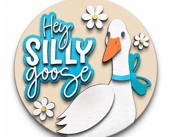 May include: A round, cream-colored sign featuring a white goose with an orange beak and a blue bow. The sign has the words "Hey Silly Goose" in blue and white lettering, with white daisy accents.