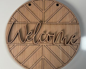 Pode incluir: Uma placa redonda de madeira com um padrão geométrico e a palavra "Bem-vindo" em uma fonte decorativa.