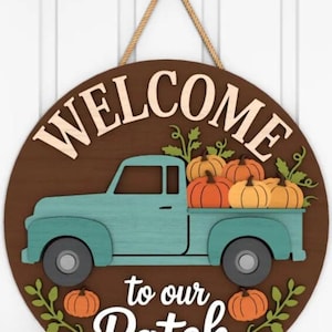 May include: Round wooden sign with a brown background and the words "WELCOME to our Patch". A teal truck carries pumpkins. The sign is decorated with pumpkins and green leaves. It hangs from a rope.