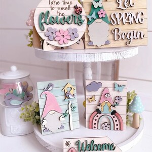 May include: Four wooden signs with a gnome theme. The signs feature a gnome with a pink hat, a rainbow, flowers, birds, and the text "Take time to smell the flowers", "Let Spring Begin", "Welcome to our Gnome", and a birdhouse with a bird.