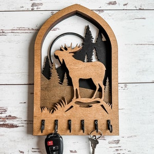May include: Wooden key holder with a moose silhouette and a forest scene. The key holder has five hooks for hanging keys.