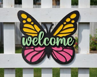 May include: A wooden butterfly-shaped welcome sign with yellow and black wings, pink lower wings, and green lettering. The sign is mounted on a white picket fence, perfect for outdoor decor.