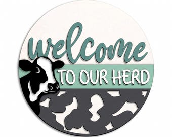 May include: A round wooden sign with the words "welcome to our herd" in teal and white. A black and white cow graphic is on the left side. The bottom half of the sign has a cowhide pattern.