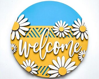 May include: Round wooden welcome sign with a blue and yellow background, daisy accents, and the word "welcome" in white script. The sign has a layered design with cut-out details.