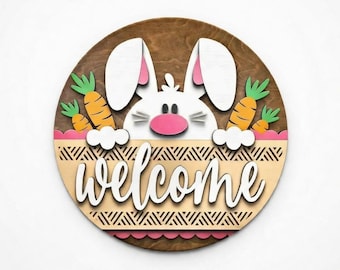 May include: A round wooden welcome sign featuring a white bunny with pink inner ears and nose, peeking over a basket with carrots. The word "welcome" is written in white script across the sign. The sign has a brown background.