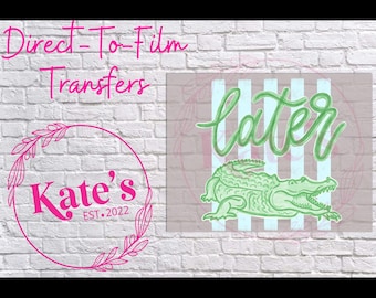 May include: Direct-To-Film Transfers design featuring the word "later" in green script above a green alligator illustration. The design is set against a striped background and a white brick wall. A pink logo with text is also present.