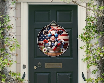 May include: A round wooden door hanger featuring a cow wearing star-shaped sunglasses and a red, white, and blue bandana. The cow is in a metal bucket with an American flag background and sunflowers. The door is dark green.