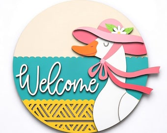 May include: A round wooden welcome sign featuring a cartoon goose wearing a pink hat and bow. The sign has a teal and yellow patterned border, with the word "Welcome" in white script. The background is a light beige color.
