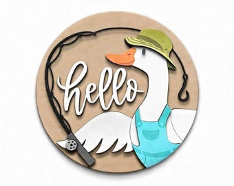 May include: A round wooden sign featuring a cartoon goose wearing a green hat and blue overalls, holding a fishing rod. The word "hello" is written in white script. The goose has an orange beak and is set against a light brown background.