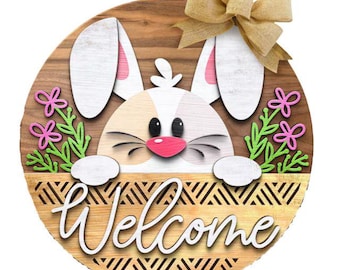May include: A wooden Easter-themed round sign featuring a bunny peeking over a basket with the word "Welcome" in cursive. The bunny has white ears with pink insides, a pink nose, and black eyes. A burlap bow and pink flowers complete the design.