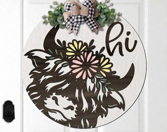May include: Round wooden door hanger featuring a Highland cow design with the word "hi". The cow has a floral crown with yellow and pink flowers. The door hanger is decorated with a buffalo check bow and greenery.