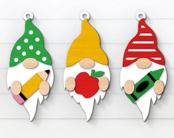 May include: Three wooden gnome ornaments with different coloured hats and school-themed accessories. The gnome on the left has a green hat with white polka dots and is holding a yellow pencil. The gnome in the middle has a yellow hat and is holding a red apple. The gnome on the right has a red and white striped hat and is holding a green crayon.
