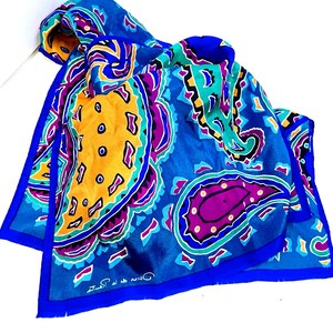 May include: A square scarf featuring a vibrant paisley pattern in shades of royal blue, bright yellow, purple, and teal. The fabric displays large, stylized teardrop shapes with black outlines and a solid blue border. The text Oscar de la Renta is printed.