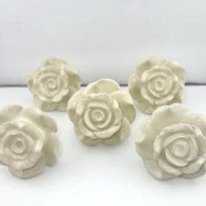 May include: Five off-white ceramic rose-shaped cabinet knobs. Each knob features detailed petals and a central spiral design. The knobs are arranged against a plain white background, showcasing their delicate floral form.