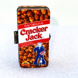 May include: A rectangular, hinged lighter featuring a Cracker Jack design. The lighter's front displays the Cracker Jack logo in red and white, with a caramel popcorn and peanut background. The design includes a sailor and his dog.