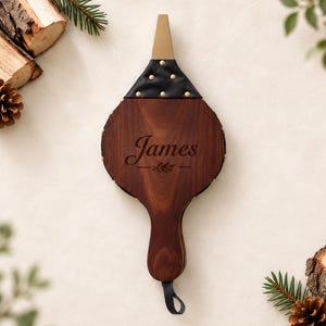 Puede incluir: Un fuelle de chimenea de madera marrón oscuro con una sección de boquilla de cuero negro y tachuelas metálicas doradas. El centro de la madera está grabado con el nombre James sobre un pequeño gráfico de hoja. Un lazo negro cuelga del mango.