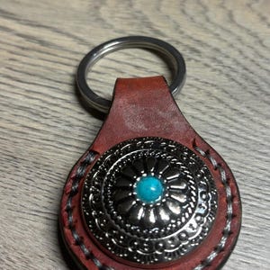 May include: A leather keychain with a decorative silver-tone metal medallion and a turquoise stone center. The keychain has a silver ring and is stitched with white thread. The leather is a reddish-brown color.