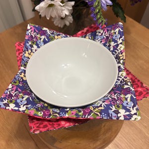 May include: A white ceramic bowl sits inside a square fabric bowl cosy. The cosy features a floral pattern with shades of purple, blue, green, and white on the interior, while the exterior displays a vibrant pink and red animal print pattern.