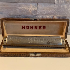 May include: A vintage Hohner Goliat harmonica in a wooden case. The harmonica has a silver finish with engraved details and the words "Goliat Made in Germany" on the front. The case is brown with a textured finish and has a brass latch.