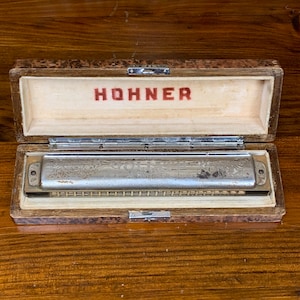 May include: A vintage Hohner harmonica in a wooden case. The harmonica has a silver finish and is decorated with floral engravings. The case is brown with a wood grain pattern and has a metal latch.