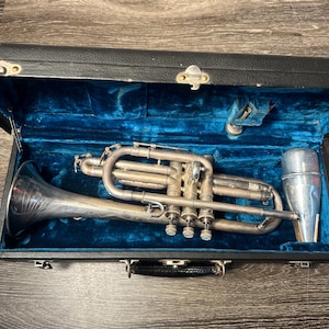 May include: A silver-colored cornet in an open black case with a blue velvet interior. The cornet has three valves and a detachable mouthpiece. The case is rectangular with a handle and a clasp.