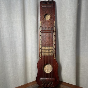 May include: A vintage, reddish-brown harp guitar with gold accents. The instrument features a round sound hole and a series of strings. The body has a unique shape, and the headstock has tuning pegs. The words "Harp Guitar" are visible on the body.