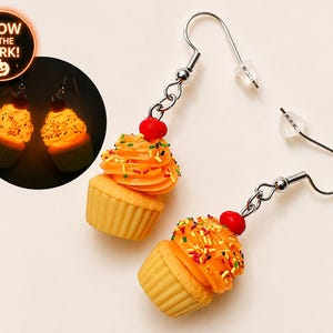 May include: A pair of dangle earrings featuring miniature cupcake charms with tan liners, orange frosting, multi-colored sprinkles, and a red cherry on top. The earrings include a circular inset showing the cupcakes glowing in the dark with the text GLOW IN THE DARK.