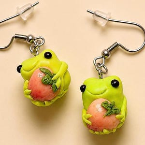 May include: A pair of drop earrings featuring small, lime green frogs with black eyes. Each frog is holding a round, pinkish-orange fruit with a small green leaf attached. The charms are suspended from silver-toned metal fishhook ear wires.