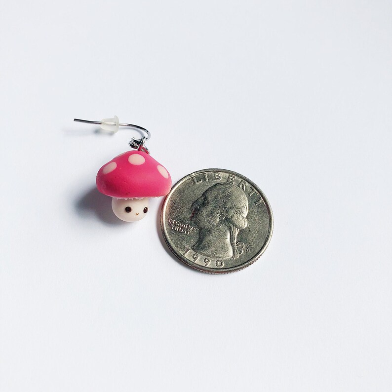 May include: A pink and white mushroom with a cute face, hanging from a silver earring hook. The mushroom is about the size of a US quarter.