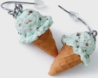May include: A pair of dangle earrings shaped like mint chocolate chip ice cream cones. Each earring features a textured light green scoop with brown flecks, sitting atop a tan waffle cone, attached to a silver-toned metal hook with a clear backing.